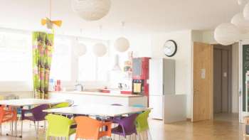 Heller Gemeinschaftsraum im müze Familienzentrum mit bunten Stühlen, runden Hängelampen und einer modernen Küche im Hintergrund.