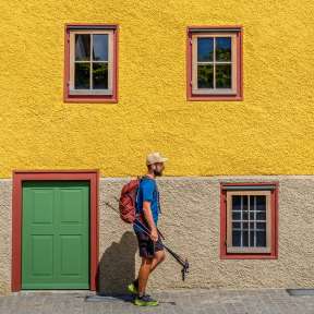 Wanderer vor dem Landweibel-Huus in Schaan mit gelber Fassade, grüner Tür und roten Fensterrahmen – ein Highlight am Liechtenstein-Weg.