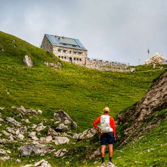 Wanderer nähert sich der Pfälzerhütte auf einem grünen Hang in Liechtenstein, eingerahmt von Felsen und Wiesen