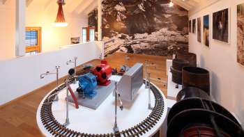 Modell einer Turbine auf einer Ausstellungsplattform im Lawena Museum, im Hintergrund historische Schwarzweißaufnahmen.