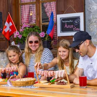 Familie sitzt auf einer sonnigen Alpe in Liechtenstein und geniesst eine traditionelle Jause mit regionalen Spezialitäten und Bergpanorama