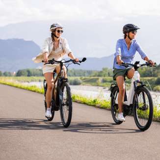 Zwei Frauen auf E-Bikes am Rheinradweg bei Liechtenstein, mit Fluss, grüner Landschaft und Bergen im Hintergrund