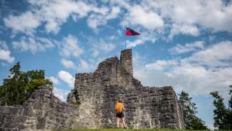 Wanderin vor der historischen Burgruine Schellenberg mit wehender Liechtenstein-Flagge unter blauem Himmel
