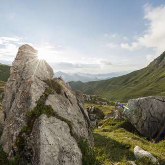Sonnenaufgang hinter Felsformationen im grünen Naaftal in Liechtenstein, umgeben von sanften Hügeln und Berglandschaft