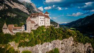 Die imposante Burg Gutenberg thront auf einem Felsen in Balzers, umgeben von grünen Hängen und majestätischer Alpenkulisse bei blauem Himmel