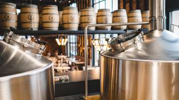 Blick in eine moderne Brauerei mit glänzenden Edelstahltanks und gestapelten Holzfässern im Hintergrund.