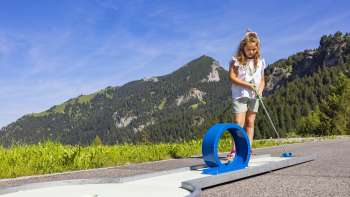 Mädchen konzentriert sich beim Minigolfspielen auf eine blaue Looping-Bahn in alpiner Kulisse