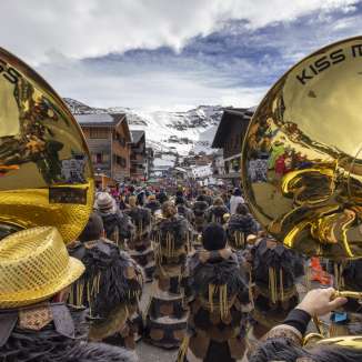 Blick auf Guggenmusik-Gruppe mit goldenen Instrumenten beim Fasnachtsumzug in Malbun vor verschneiter Bergkulisse.