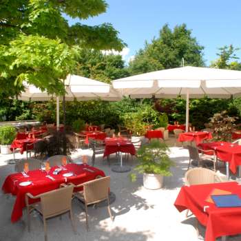 Außenansicht der Terrasse mit roten Tischdecken, umgeben von Bäumen und Sonnenschirmen.