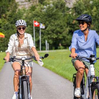 Zwei Frauen auf E-Bikes bei sommerlicher Tour mit Flaggen von Liechtenstein, Schweiz und Österreich im Hintergrund