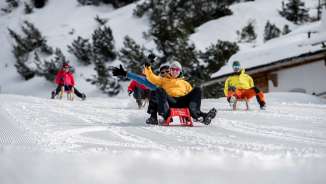 Gruppe fröhlicher Rodler auf einer präparierten Piste in Malbun, umgeben von verschneiten Bäumen und alpiner Winterlandschaft