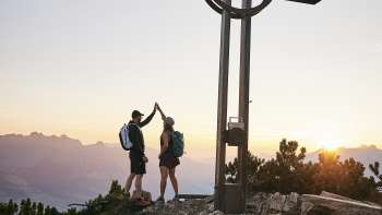Zwei Wanderer geben sich ein High-Five am Gipfelkreuz beim Alpspitz bei Sonnenaufgang mit Bergpanorama im Hintergrund.