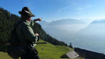 Ein Mann in traditioneller Kleidung zeigt von einer Bergwiese aus ins Rheintal.