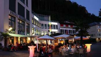 Abendliche Stimmung vor der Brasserie Burg in Vaduz mit beleuchteter Terrasse und Gästen unter Sonnenschirmen.