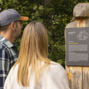 Zwei Personen lesen eine Infotafel mit Sage am WalserSagenWeg, auf einer geschnitzten Holzfigur