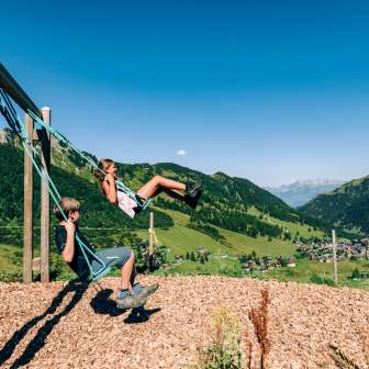 Zwei Kinder schaukeln auf einem Bergspielplatz mit Blick über das Sommerpanorama von Malbun in Liechtenstein