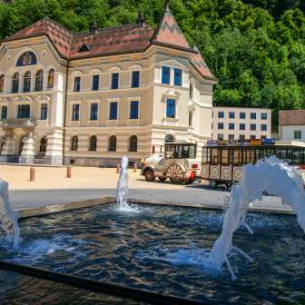 Blick auf das Regierungsgebäude in Vaduz mit vorbeifahrendem Citytrain und Wasserfontänen im Vordergrund