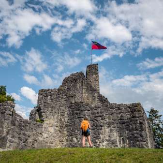 Wanderin vor der historischen Burgruine Schellenberg mit wehender Liechtenstein-Flagge unter blauem Himmel