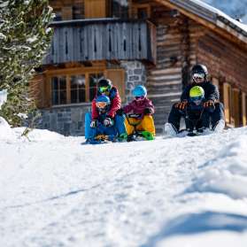 Zwei Erwachsene und drei Kinder sitzen auf Schlitten und starten gemeinsam eine Abfahrt vor einer verschneiten Berghütte in Malbun