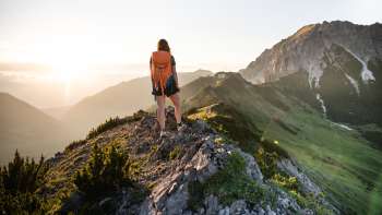 Wanderin mit Rucksack steht auf einem Bergkamm in Liechtenstein und blickt in die aufgehende Sonne.