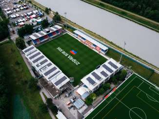 Luftaufnahme des Rheinpark Stadions in Vaduz mit „Hoi aus Vaduz“-Schriftzug auf dem Spielfeld und dem Rhein im Hintergrund