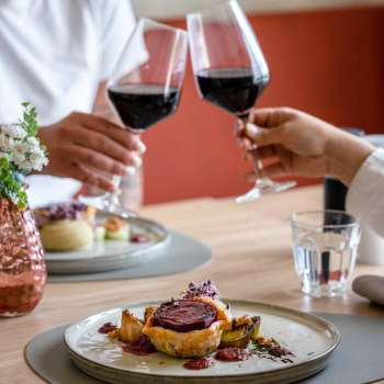 Zwei Personen stoßen mit Rotwein an, im Vordergrund ein liebevoll angerichtetes Hauptgericht