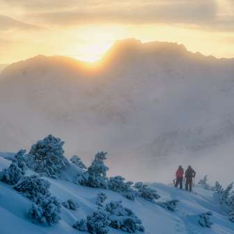 Zwei Personen, darunter Tina Weirather, wandern durch verschneite Winterlandschaft bei Sonnenuntergang in den Bergen Liechtensteins.
