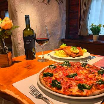 Nahaufnahme einer Pizza mit Tomaten, Käse und Basilikum auf einem Holztisch, im Hintergrund ein Glas Rotwein.