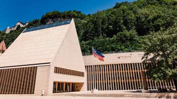 Modernes Landtagsgebäude in Vaduz mit spitzem Giebel, davor die Liechtenstein-Flagge und umgeben von grüner Hügelkulisse.