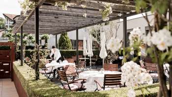 Eine elegante Terrasse mit weißen Tischen, gedeckt mit Servietten und Gläsern unter einem Holzpavillon, umgeben von üppigem Grün.