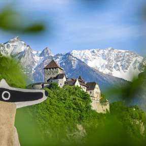 Blick auf das Schloss Vaduz mit Detektiv-Trail-Maskottchen im Vordergrund und verschneiten Alpen im Hintergrund.