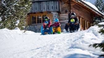 Rodeln mit Kindern in Malbun Zwei Erwachsene und drei Kinder sitzen auf Schlitten und starten gemeinsam eine Abfahrt vor einer verschneiten Berghütte in Malbun