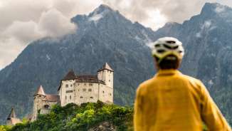 Radfahrer mit Helm blickt auf die imposante Burg Gutenberg vor dramatischer Bergkulisse in Balzers – Kulturtour mit dem Fahrrad in Liechtenstein