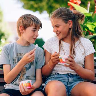 Zwei Kinder sitzen auf einer Parkbank in Liechtenstein und essen Eis – lachende Gesichter, Blumen und Sommerstimmung im Grünen