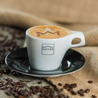 Cappuccino mit Krone im Schaum in einer Tasse von Demmel Kaffee, umgeben von Kaffeebohnen auf Holz und Jutesack – Genuss in Liechtenstein