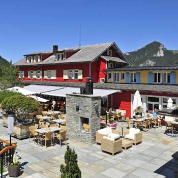 Außenansicht des Restaurants bei Sonnenschein, mit roter Fassade und bergigem Panorama.