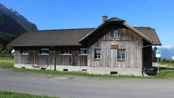 Historisches Holzgebäude der Alten Säge in Schaanwald vor Alpenkulisse bei blauem Himmel.