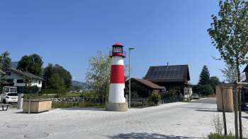 Kleiner rot-weißer Leuchtturm Moby auf einem Platz in Ruggell bei sonnigem Wetter, mit traditionellen Gebäuden im Hintergrund.