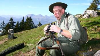 Ein Jäger mit Fernglas sitzt auf einer Wiese vor alpiner Bergkulisse.