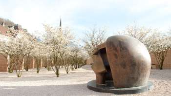 Bronzeskulptur „Figure in a Shelter“ von Henry Moore im Skulpturenpark Vaduz – umgeben von blühenden Bäumen im Frühling.