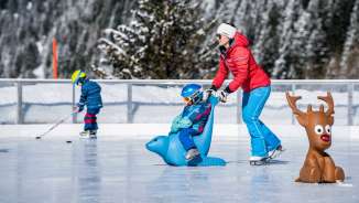 Mutter schiebt Kind auf einem Eislauf-Hilfspinguin über den Eisplatz in Malbun, im Hintergrund ein Kind mit Eishockeyschläger