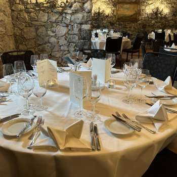 Elegant gedeckter runder Tisch mit weißen Tischdecken, Servietten und Speisekarten vor einer Steinwand.