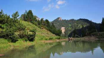 Ein See an dem zwei Wanderer sitzen, umgeben von grünen wiesen und Wald, im Hintergrund sieht man die Liechtensteiner Alpen.
