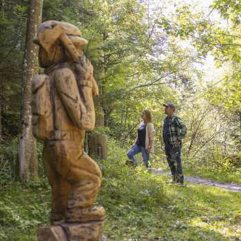 Zwei Wanderer entdecken eine geschnitzte Figur auf dem WalserSagenWeg inmitten eines lichtdurchfluteten Waldes
