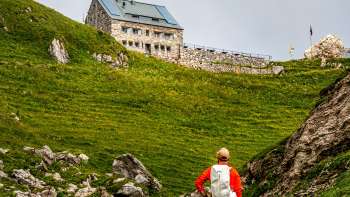 Wanderer mit rotem Rucksack blickt auf die Pfälzerhütte inmitten grüner Berglandschaft.