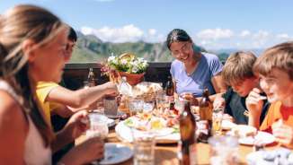 Familie beim gemeinsamen Mittagessen auf einer Sonnenterrasse in Sarreis, umgeben von Bergen und regionalen Spezialitäten
