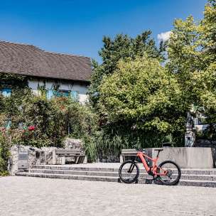 Rotes E-Bike vor historischem Gebäude in Liechtenstein, umgeben von Bäumen und Brunnen, idealer Ausgangspunkt für eine Radtour durchs Land