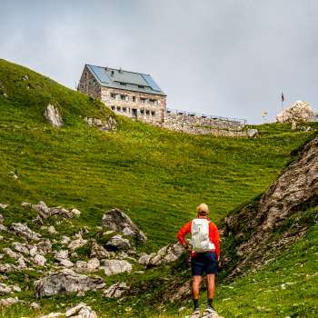 Wanderer mit rotem Rucksack blickt auf die Pfälzerhütte inmitten grüner Berglandschaft.