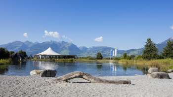 Naturnaher Badebereich mit Sand, Treibholz und Aussicht auf die umliegenden Berge