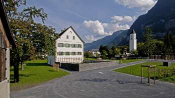 Historischer Pfarrhof mit weißer Fassade und grünen Fensterläden inmitten eines offenen Platzes, im Hintergrund die Kirche von Balzers und die umliegende Berglandschaft.